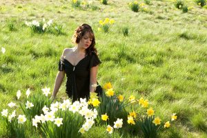 A model wearing a black dress seated in a field of daffodils.
