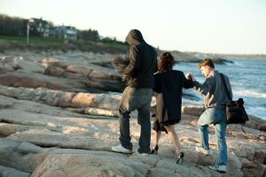 Two men help a model wearing heels off a ledge by the ocean's edge