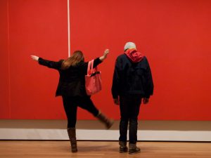 A woman dances in front of a red painting while her companion looks on.