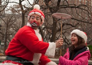 A juggling Asian Santa in Union Square helping a girl spin a plate on a stick.