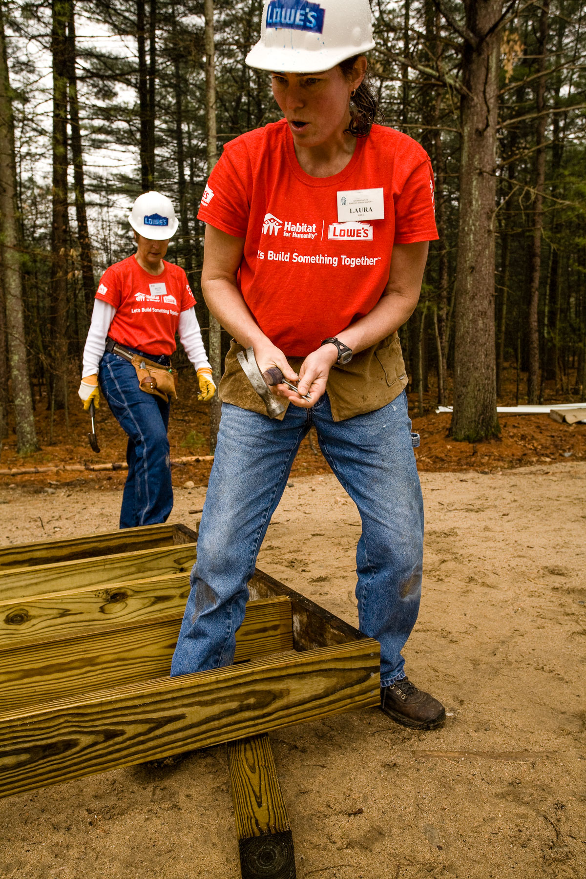 Two women in red Habitat for Humanity tee shirts and hard hats work on framing for a house