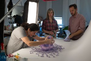 A designer arranges gift boxes and jelly beans for a shoot for a florist client as the client and graphic designer look on.