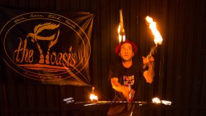 Martin Kuborn juggling flaming totrches while twirling a hula-hoop with six flaming torches around its perimeter, all while balanced on a balance board at The Floasis.