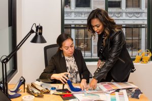 Two women work on designs for shoes, with a color swatch book and shoe samples on a desk