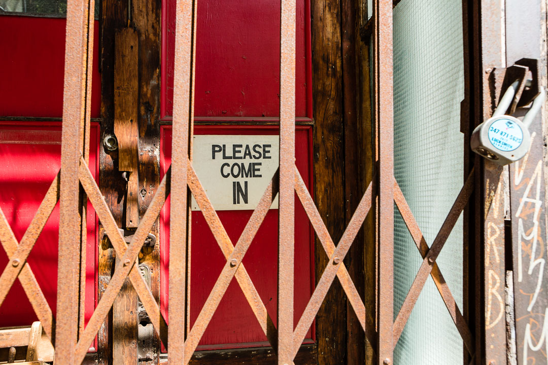 A grated shop door with the sign "Please Come In" behind the locked grate.
