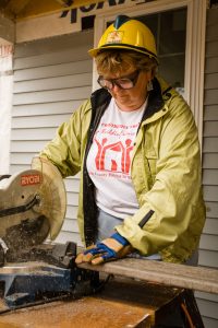 A woman in a yellow hardhat cutting a deck board for Habitat for Humanity